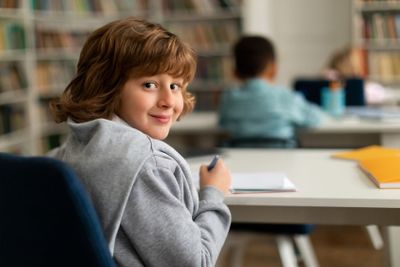 Smart male elementary school pupil, sitting at desk in...