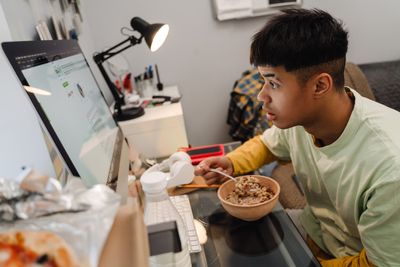 Teenage boy using desktop computer while eating...